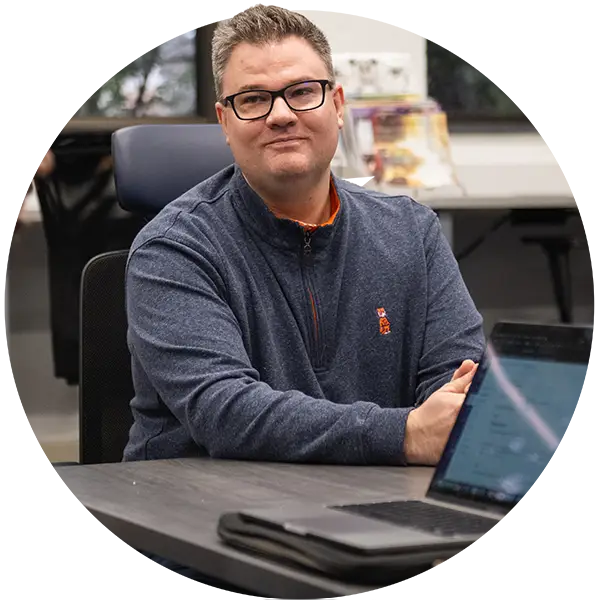 A male professor wearing glasses and a gray quarter-zip with an orange Clemson Tiger logo sits at a table with an open laptop on it.