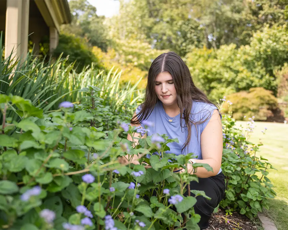 A female student wearing black pants and a lilac sleeveless top kneels down next to a collection of ornamental flowers and green plants in a botanical garden