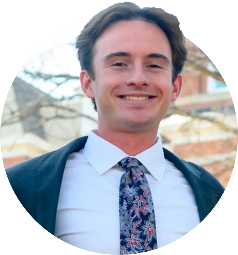 A male student in a floral tie and navy blazer poses outdoors on campus.