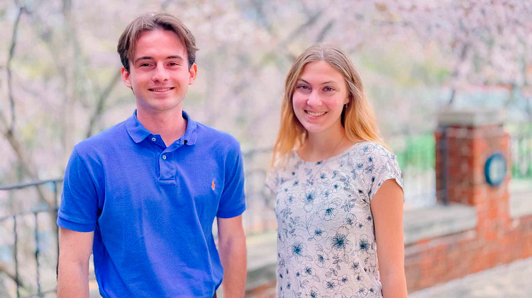 A male student wearing a blue polo and a female student wearing a white short-sleeved shirt with a blue floral pattern stand outside in front of a brick ledge and wrought-iron fencing.  