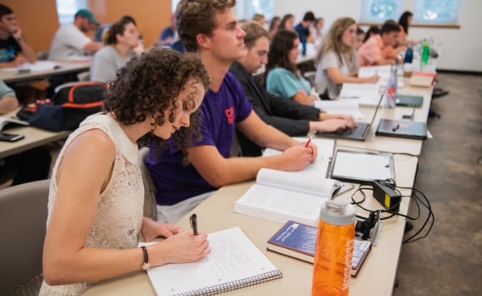 A female student takes notes while others in her class listen to the teacher at the front of the room.