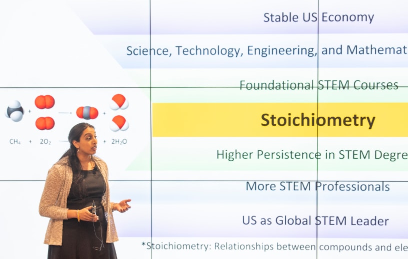 A graduate student presents her work on stoichiometry with a large screen behind her.