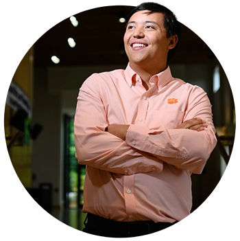 A male student wearing an orange Oxford button-down shirt with the Clemson  Tiger Paw logo and black pants stands in the lobby of the Brooks Center for the Performing Arts.