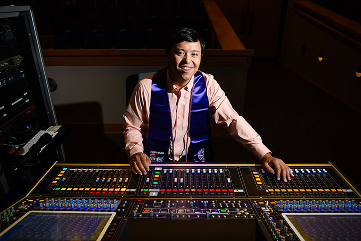 A male student wearing a long-sleeved orange Oxford shirt and a purple graduation stole sits behind a sound engineering production tool