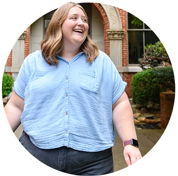 A female student wearing a light blue short-sleeved blouse and dark gray pants walks in front of a brick academic building.