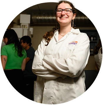 A female student wearing a white lab coat and eyeglasses poses with her arms crossed in a laboratory.