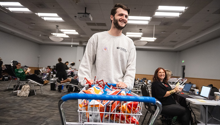 A male student in a grey The Blood Connection shirt pushes a cart full of snacks and drinks at a blood donation event