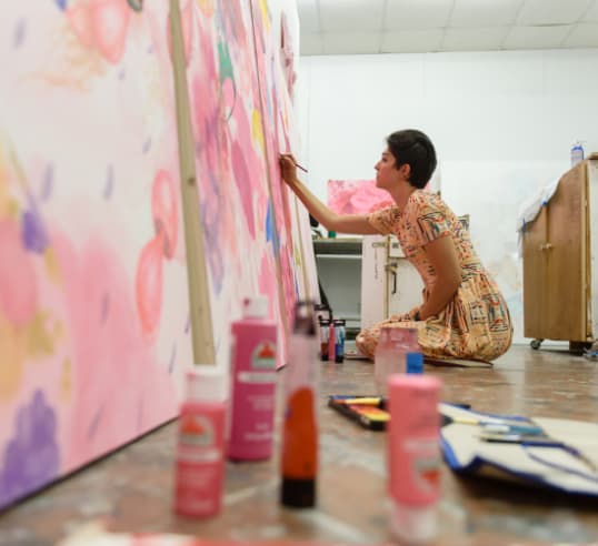 A female art student paints on a large canvas in the studio with paint bottles scattered beside her on the floor.