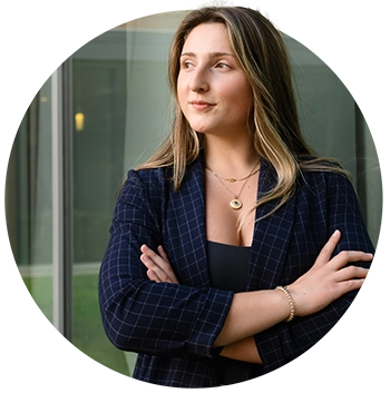 A female student wearing a navy windowpane-patterned blazer, black top and light-wash jeans poses in front of a glass wall of an academic building.