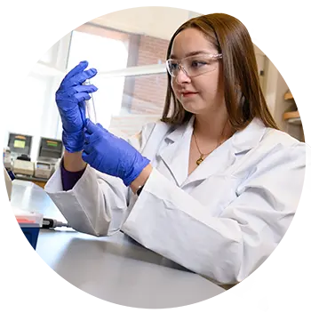 A female student wearing gloves, a white lab coat and protective eyewear looks at a graduated cylinder beaker while working in a laboratory.