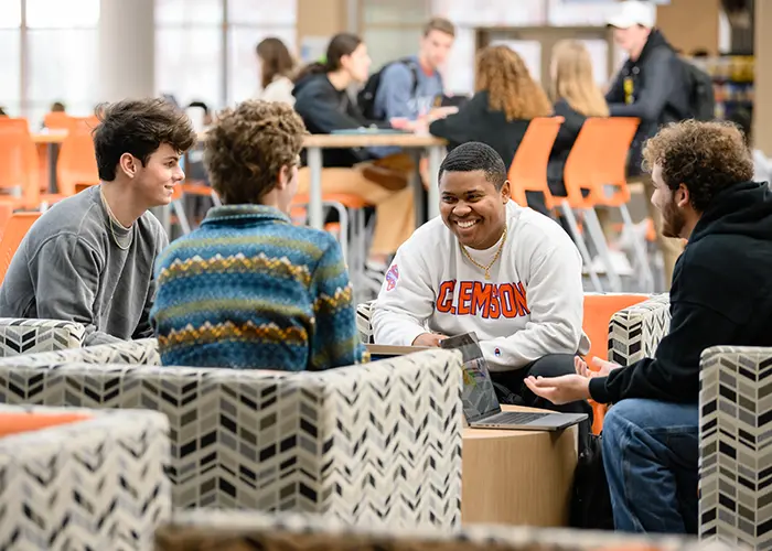 A group of male students in a student lounge area sits on patterned chairs to form a circle. An open laptop computer sits in front of one of the students.