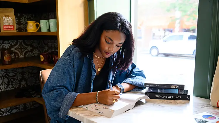 a female student sits at a table in the window of a coffee shop annotating a large book
