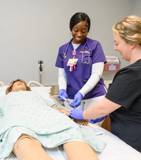A female nursing student wearing purple and white medical scrubs and a female nursing instructor wearing black scrubs stand bedside in a teaching simulation lab next to a mannequin patient lying in a hospital bed and wearing a hospital gown.