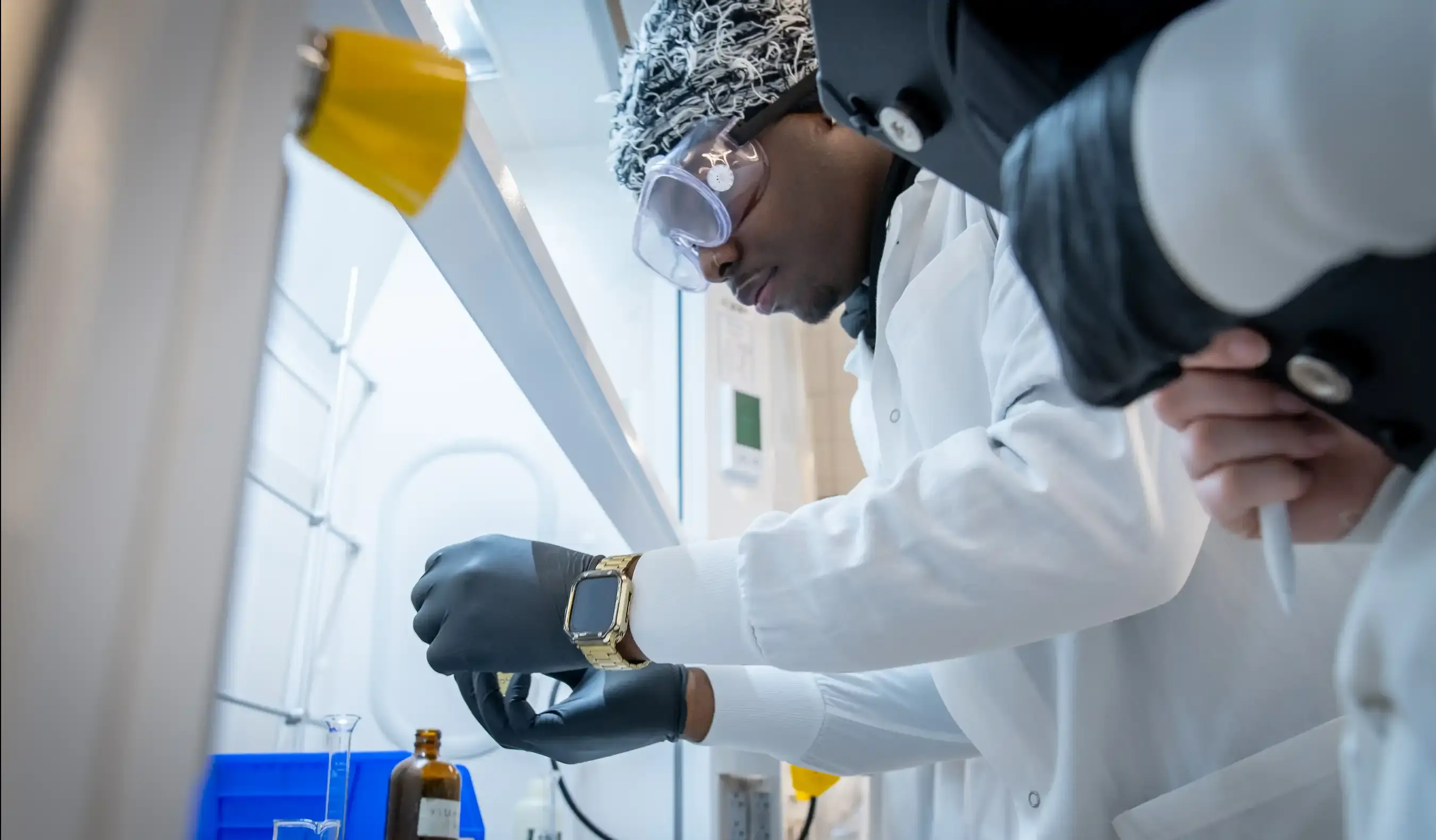 A scientist in a lab coat and gloves works with a small container under a fume hood.
