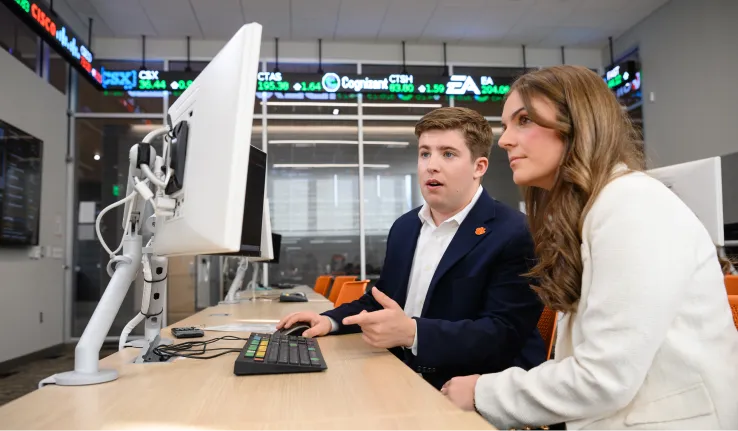 A male student wearing a navy blazer and white button-down shirt and a female student wearing a white blazer sit together at a computer in a trading room, with a stock ticker visible toward the top of the walls.