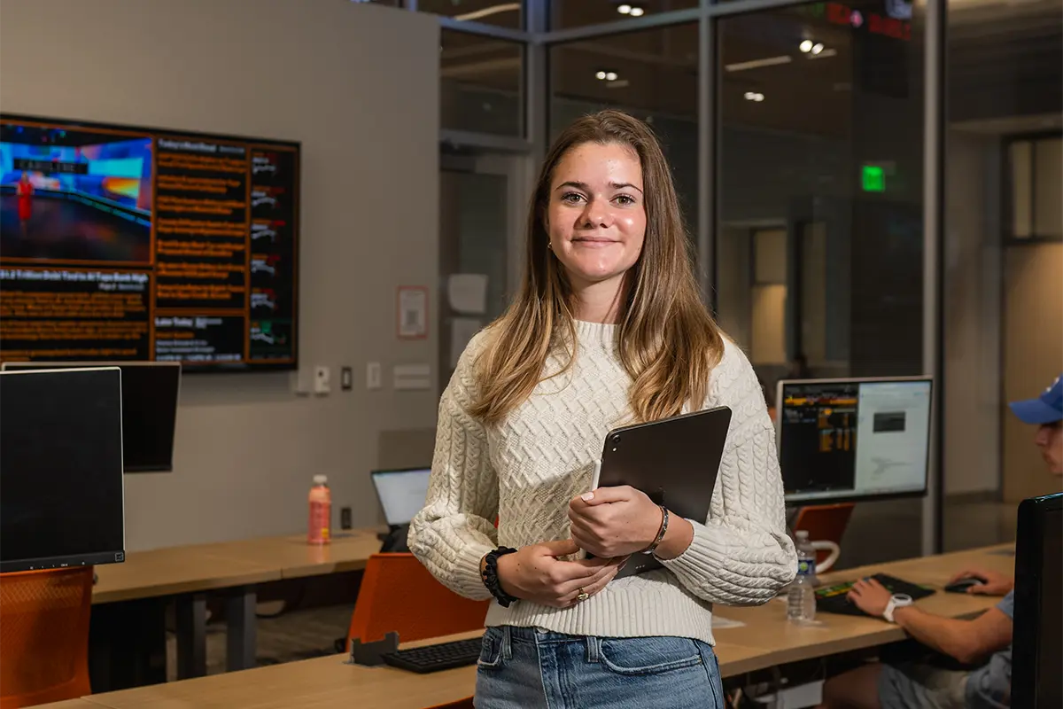 A female student wearing blue jeans and a white sweater holds an iPad while standing between tables in a classroom with a stock ticker display.