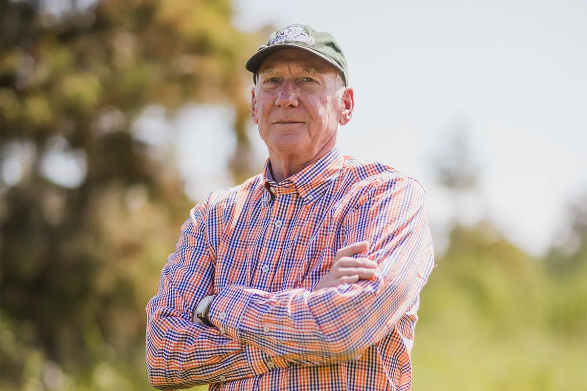A man wearing blue jeans, an orange-and-purple checkered button-down shirt and an olive green baseball cap stands among green crops in a field.