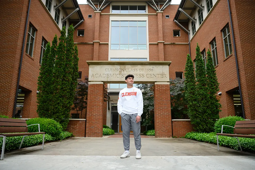 A male student wears a white sweatshirt with Clemson written in orange letters outlined in purple and gray athletic pants.