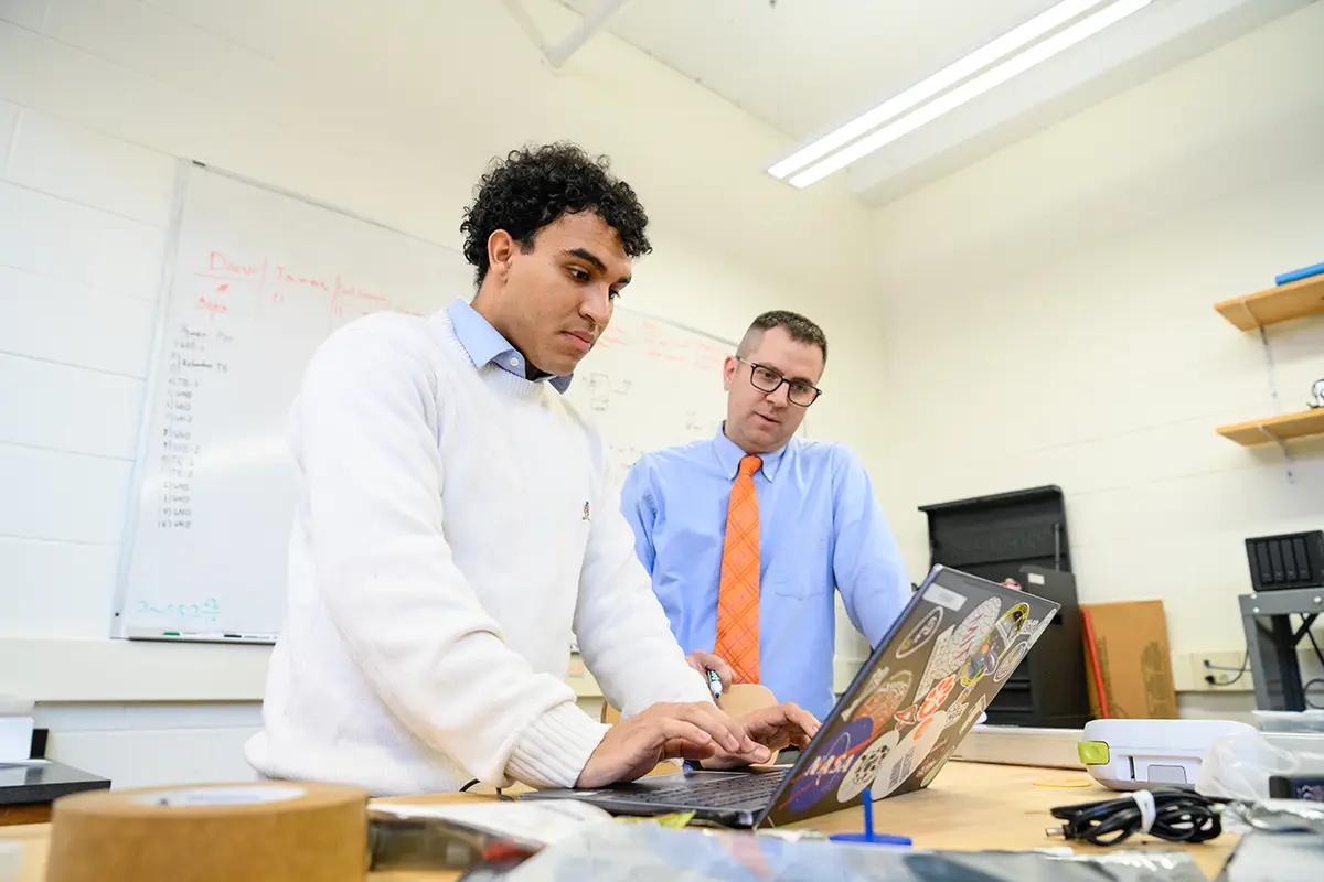 A male student wearing a cream-colored sweater over a light blue button-down shirt and a male professor wearing a light blue button-down shirt and orange tie stand in front of a laptop covered in outer space-themed stickers.