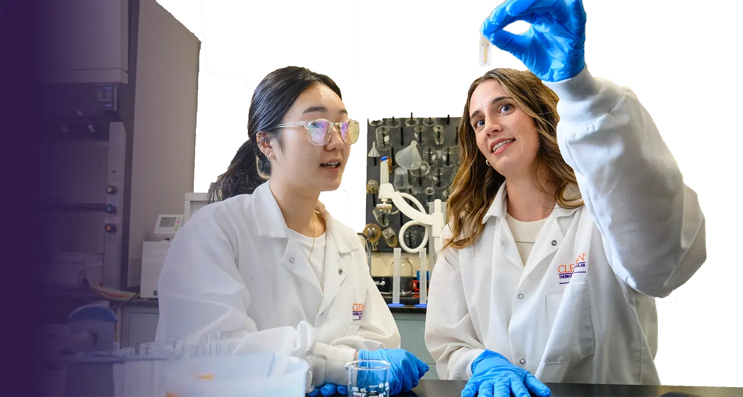 Two female students in lab coats display a test tube, highlighting their collaboration in a campus research environment.