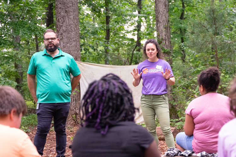 Faculty member Steph Dean and Kevin Stinehart, fourth grade teacher, address a group of his students in the middle of a forest