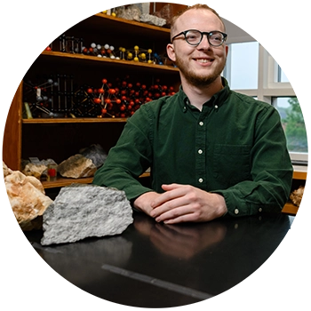 : A male student wearing a button-down dark green Oxford shirt and glasses sits in a geology laboratory with two geodes next to him on a table. 