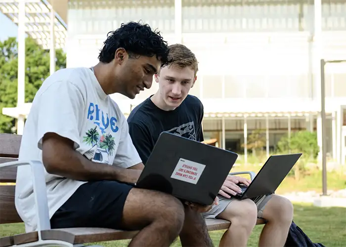 Two male students sit outside on a bench with their laptops open looking at a screen of one of the open laptops.