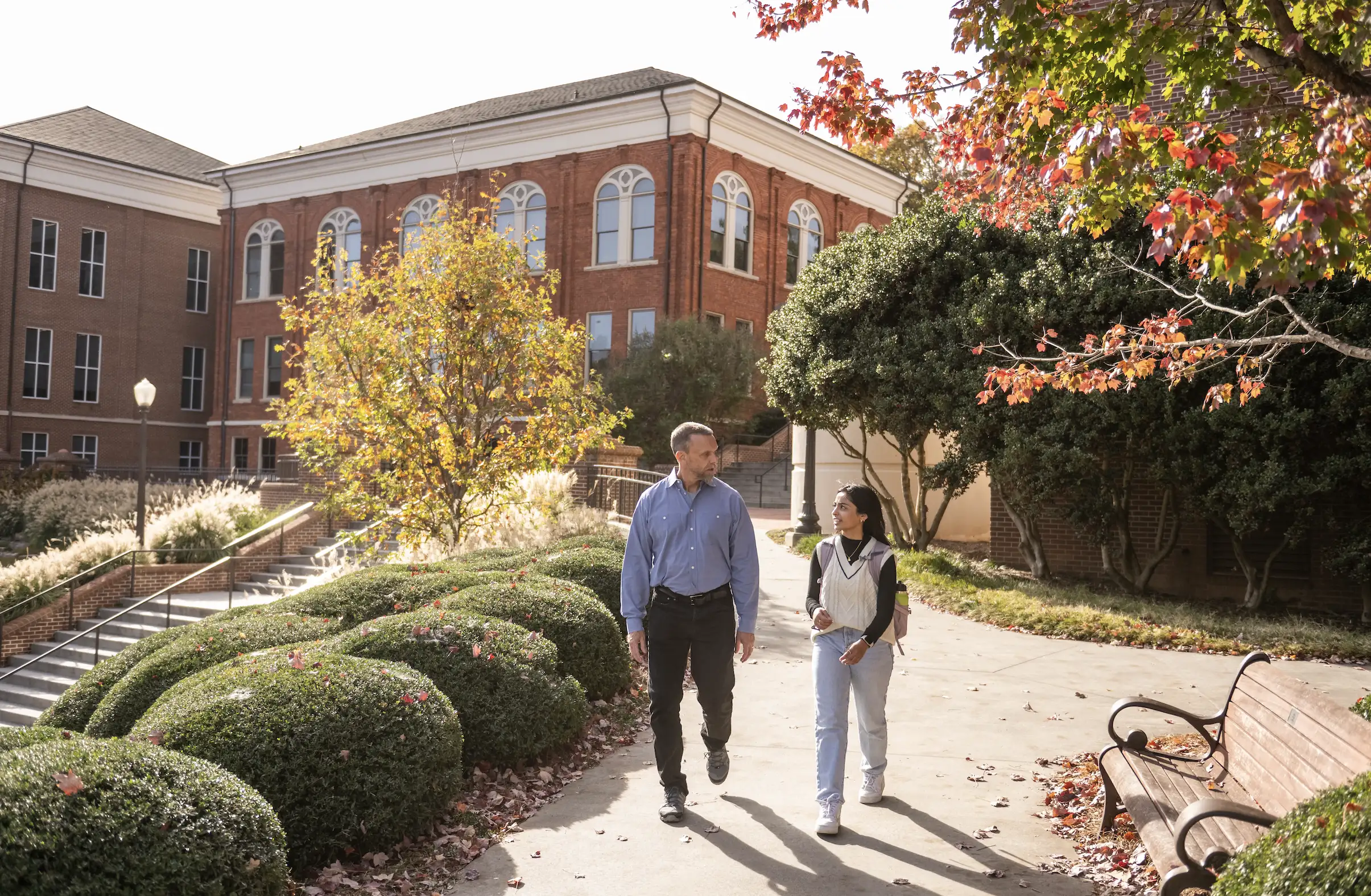 A student and her professor walk and talk on campus with an academic building, trees, and bushes in the background.