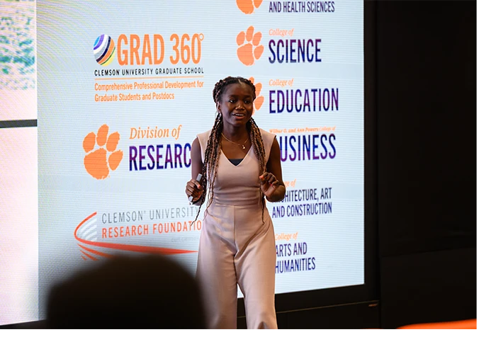 A female graduate student presents on stage with a digital screen of Clemson division and school logos behind her.