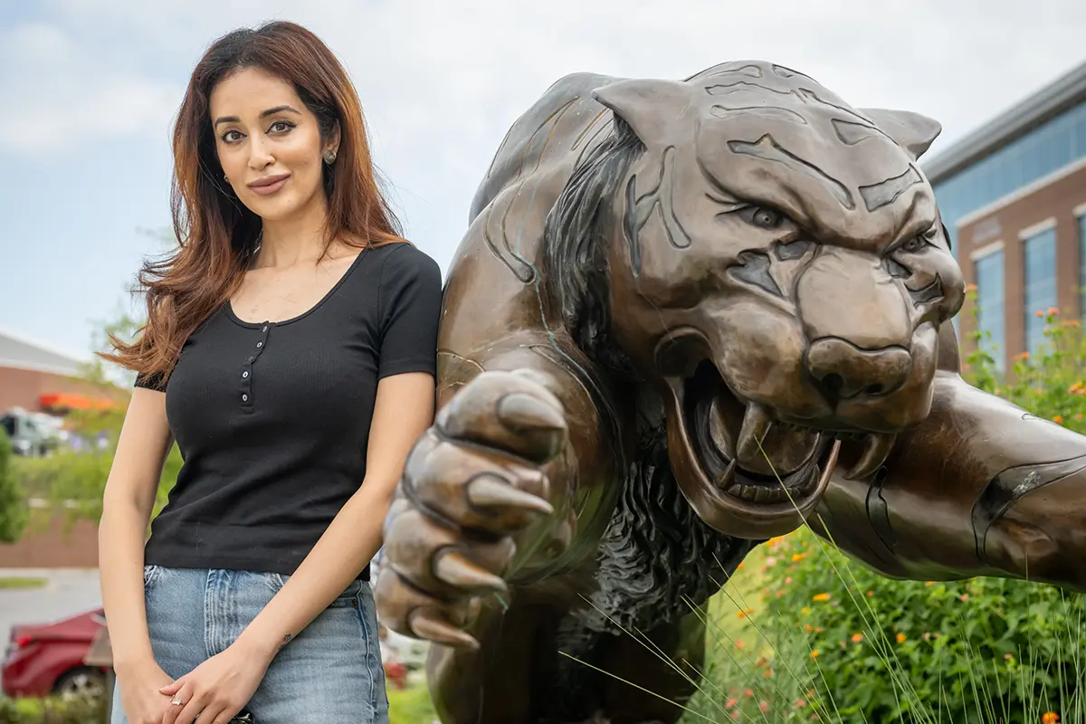 A woman wearing denim jeans and a black short-sleeved top stands next to a bronze tiger statue.