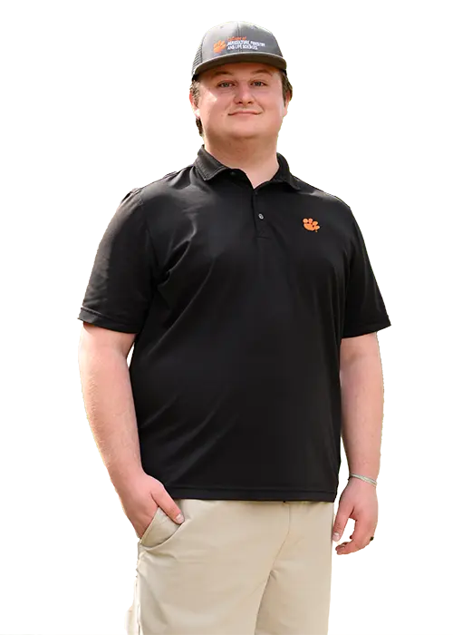 A male graduate student wearing khaki shorts, a black polo with an orange Clemson Tiger Paw and a gray baseball cap.