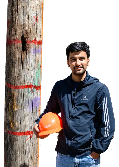 A male graduate student wearing blue jeans, work boots, a black jacket, neon yellow safety vest and orange hard hat squats down next to a timber pile and holds a small black device.