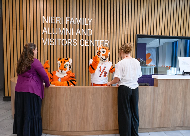 The Tiger and Cub mascots stand behind a welcome desk at the Nieri Family Alumni and Visitors Center while interacting with two female staff members.