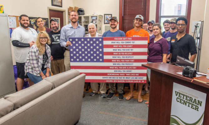 A group of student veterans holds and poses next to an American flag with patriotic slogans written on it.