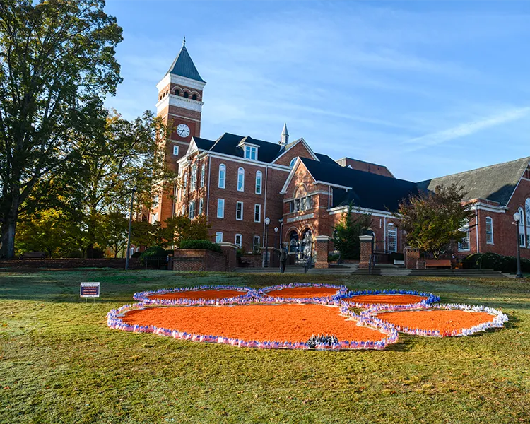 Small American flags border an orange spray-painted Tiger Paw on Bowman Field in front of the brick Tillman Hall academic building and clock tower.
