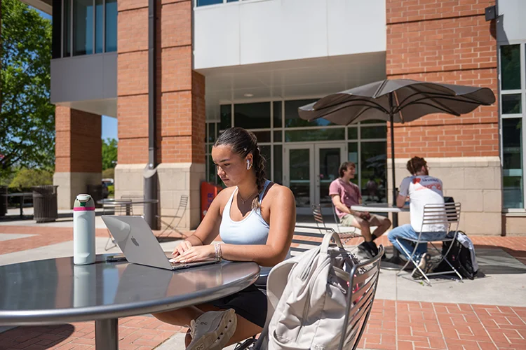 a female student works at her laptop at an outdoor table on a sunny spring day