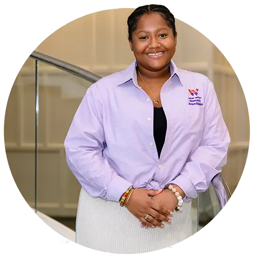 A female student wearing a cream-colored skirt, black top and light purple button-down long-sleeved shirt poses on a staircase.
