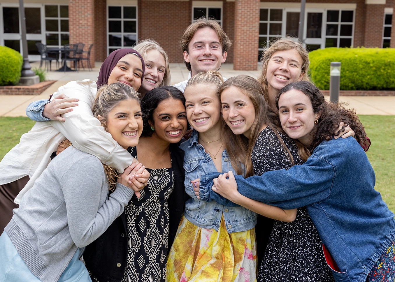 A group of nine college-age students stand closely together on a grassy lawn, smiling and hugging in front of a brick campus building with large windows and columns.