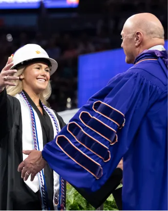 A female student wearing a white construction hard hat and a black graduation gown hugs Clemson President Jim Clements at graduation.