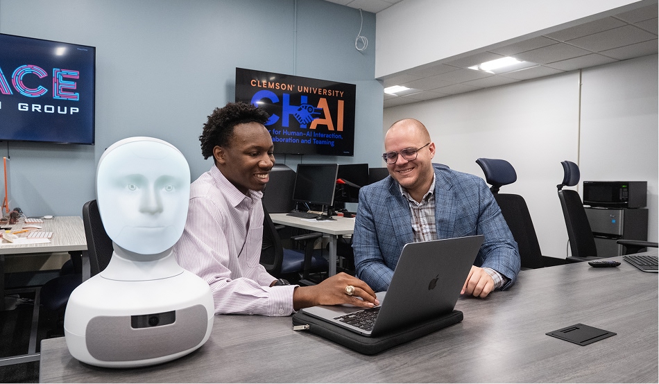 A male student and a male professor sit at a table in front of a laptop and next to an anatomical model of a human head.