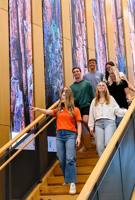 A group of students ascending stairs inside a building, engaged in conversation and carrying backpacks.