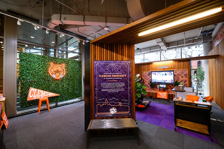 The interior of the Experience Clemson space, which includes informational signage, a green artificial hedge wall with a light-up Tiger head hung on it, and lounge space with chairs and a television mounted to a wall.
