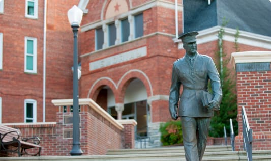 A statue of a male soldier in the foreground and a brick academic building in the background.