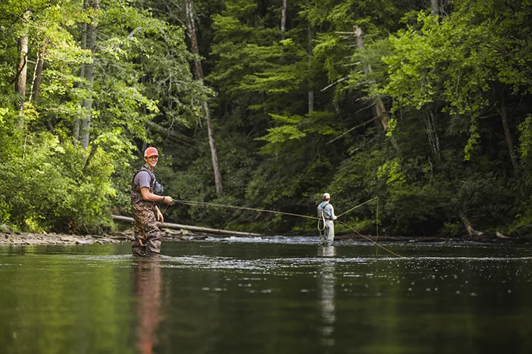 Two male students wear fishing waders and baseball caps stand in a stream holding fishing poles.