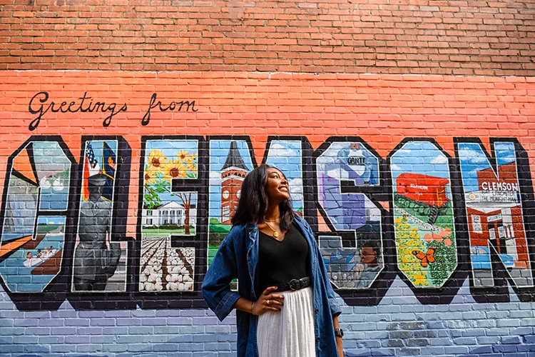 A female student in a white skirt, black T-shirt and denim oversized shirt poses with one hand on her hip in front of a colorful mural that says, Welcome to Clemson designed like a vintage postcard.