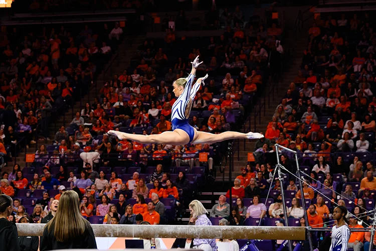 A female gymnast wearing a purple and white leotard does the splits while leaping during a balance beam routine.