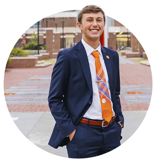 A male student wearing a navy blue suit, white shirt and purple-and-orange Clemson tie stands on a concrete and brick walkway in front of a brick-and-glass building.