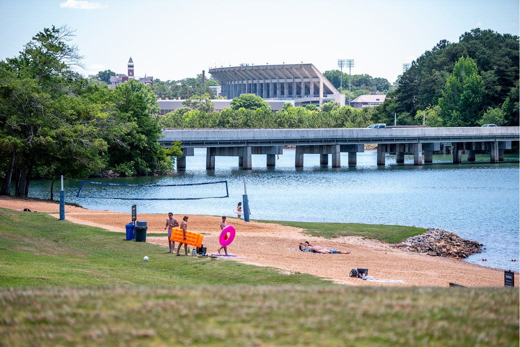 Green grass, a sandy beach volleyball court and a lake with a clock tower building and football stadium distant in the background.